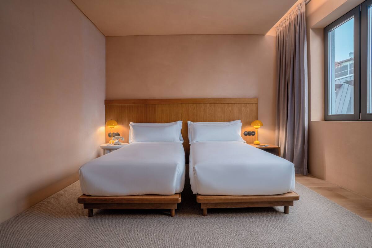 Uma cama de casal com cabeceira de madeira, duas mesas de cabeceira com luminárias e uma janela grande com cortinas.