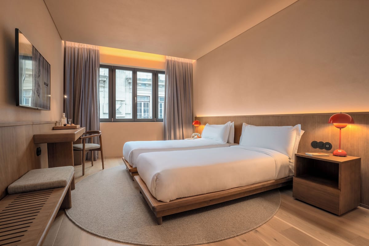 Quarto de hotel moderno com uma cama grande, mesas de cabeceira, uma escrivaninha e uma janela com cortinas.