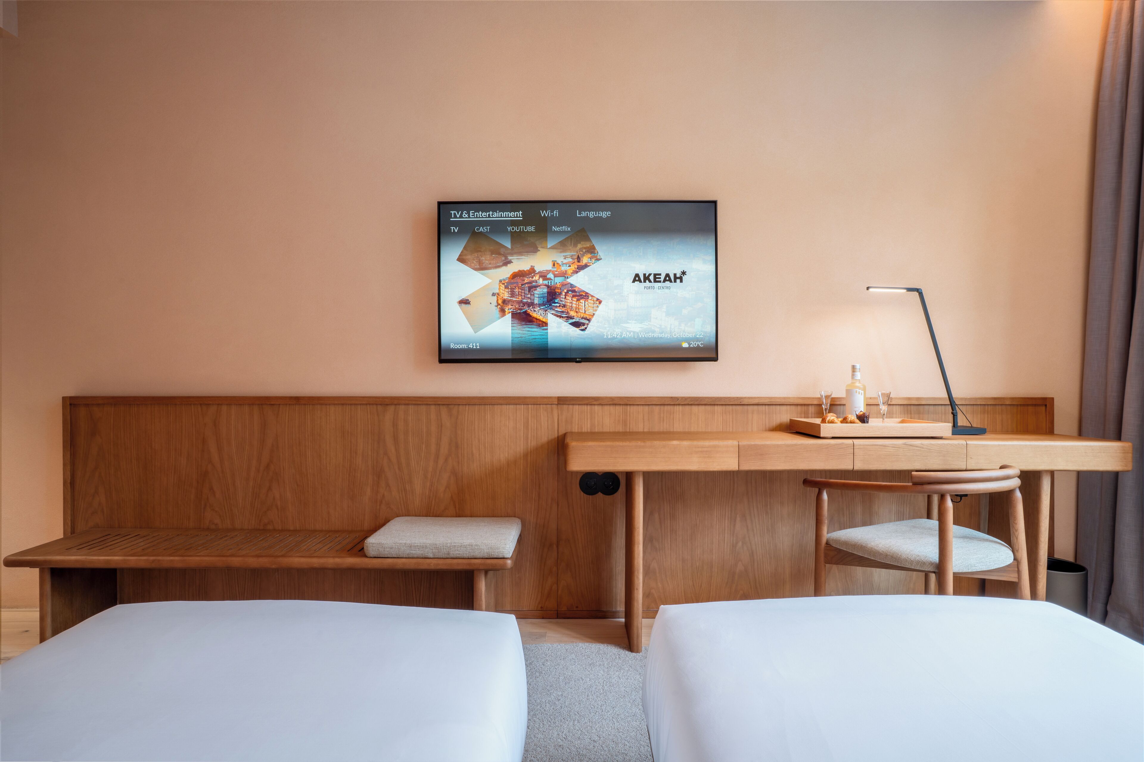 A hotel room with a bed, a desk with a laptop, a TV displaying a cityscape, and a window with curtains.