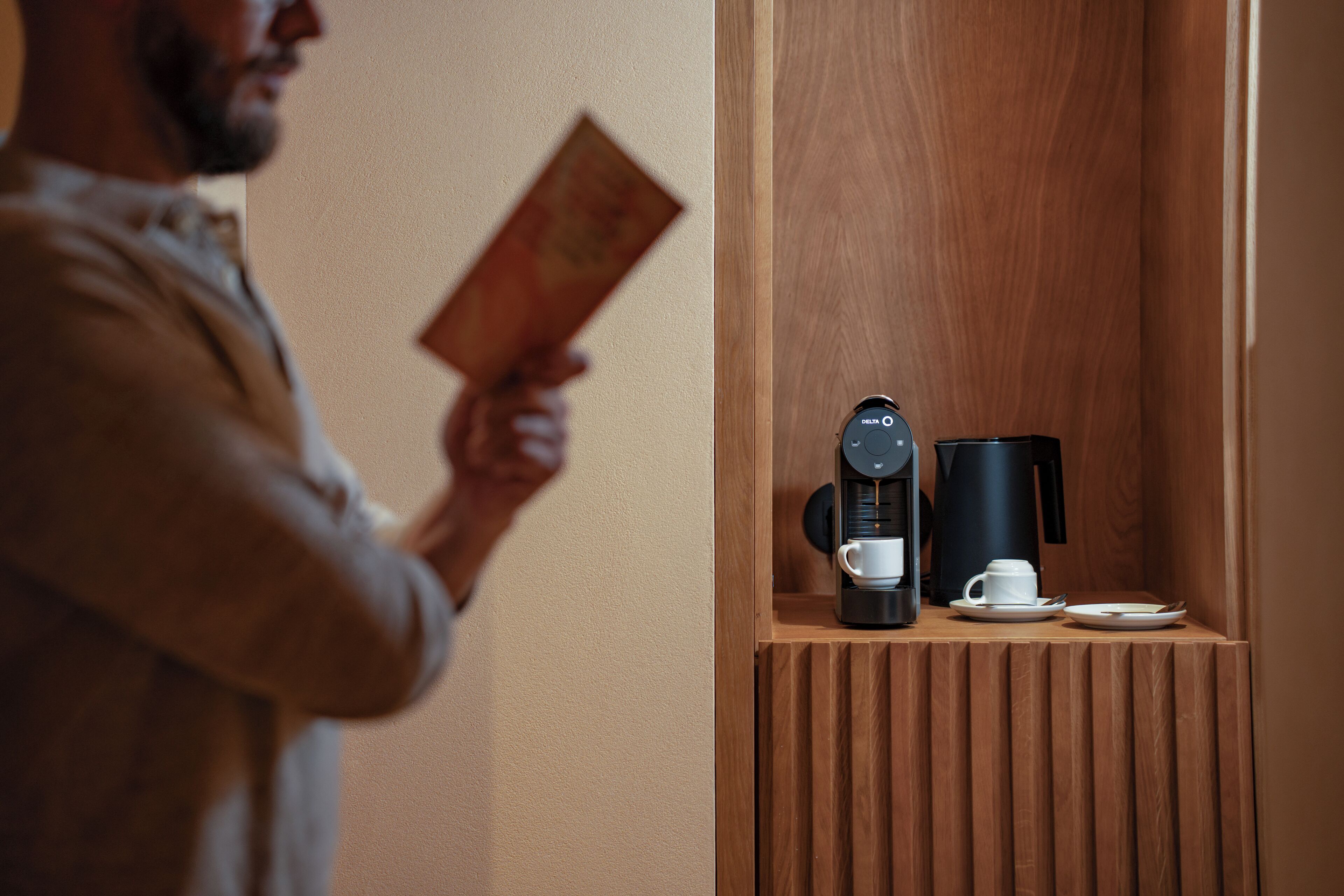 A person holding a book standing next to a wooden shelf with a coffee maker and a cup.