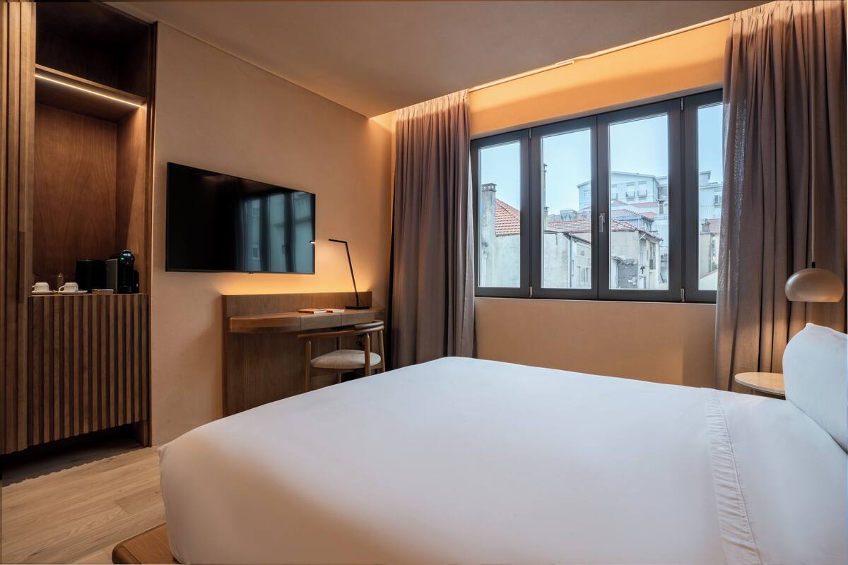 Habitación de hotel con una cama grande, un escritorio con silla, un televisor de pantalla plana montado en la pared y una ventana con cortinas.