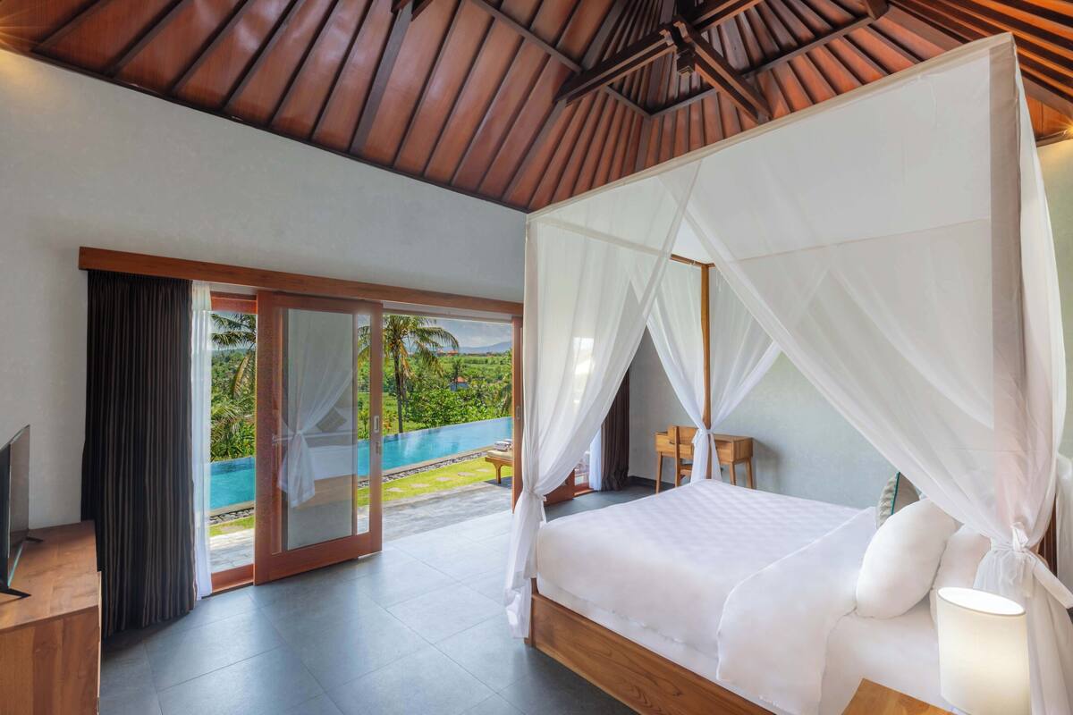 A bedroom with a bed, a TV, and a view of a pool and greenery.