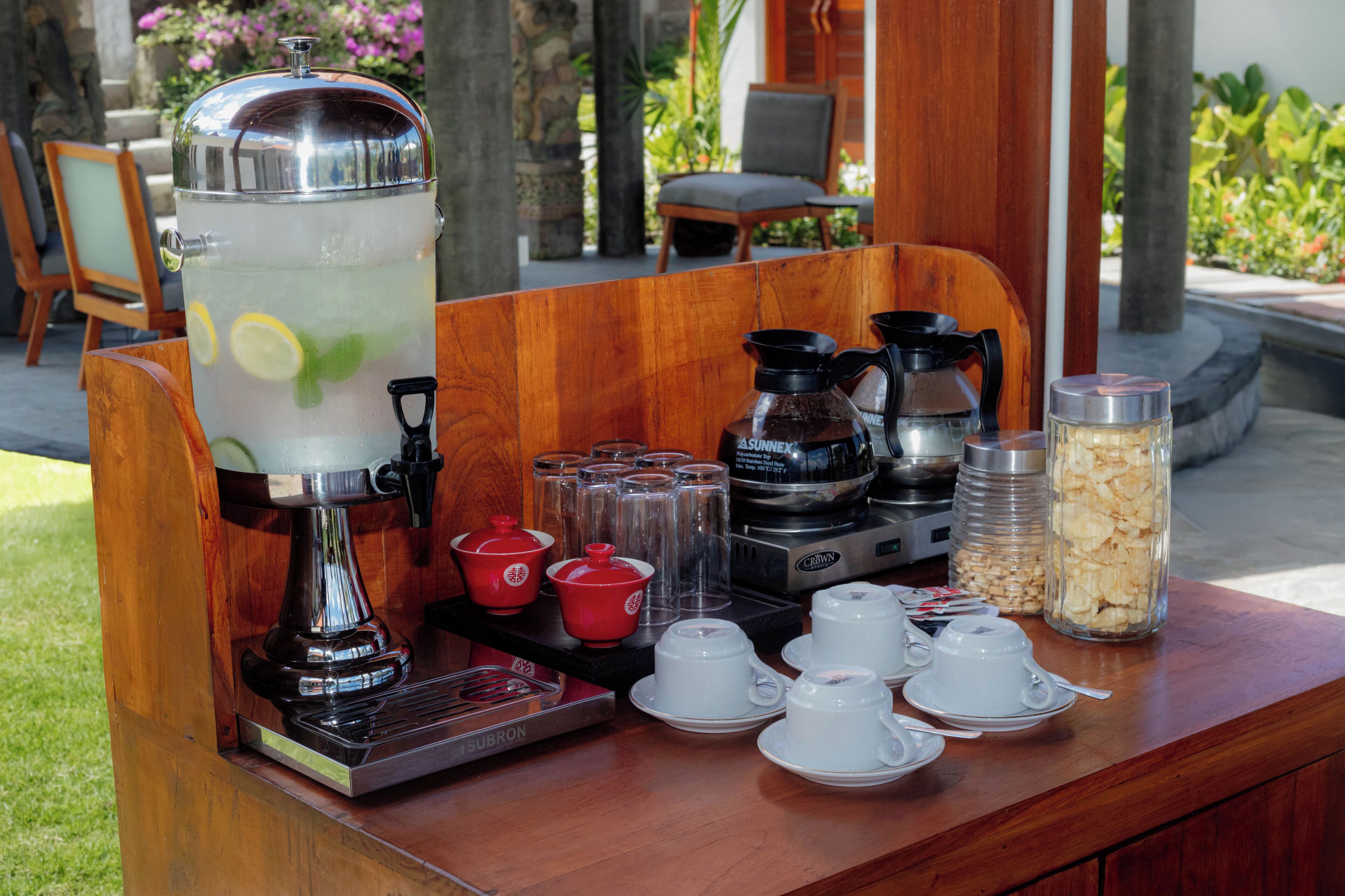 A breakfast setup with a coffee maker, a water dispenser, and various cups and containers on a wooden table.