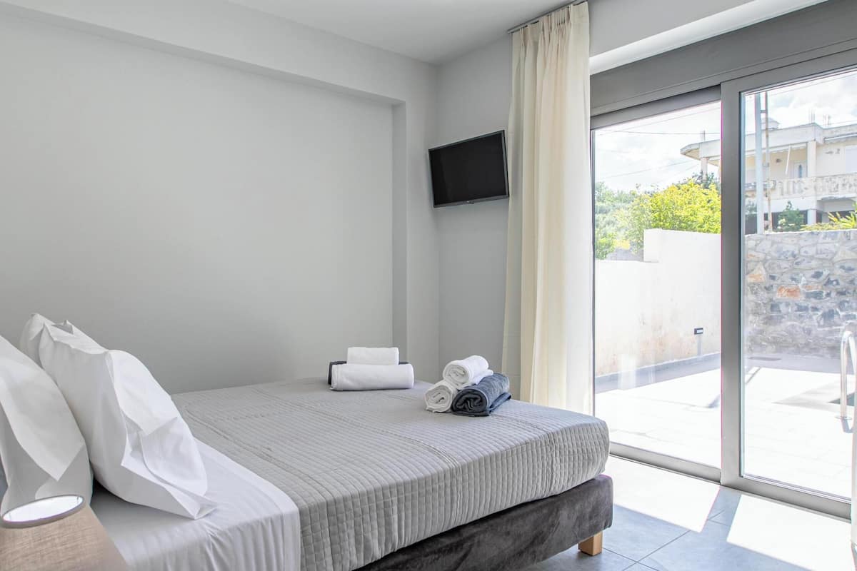 Una habitación de hotel moderna con cama, mesita de noche de madera, televisor de pantalla plana y puerta corredera de vidrio al exterior.