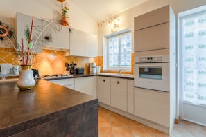 A modern kitchen with a central island, built-in oven, and a window with patterned curtains. - Holiday Cottage "Mas de la Ravine chez Juju" with Shared Pool, Wi-Fi & Air Conditioning (Laval-Saint-Roman)