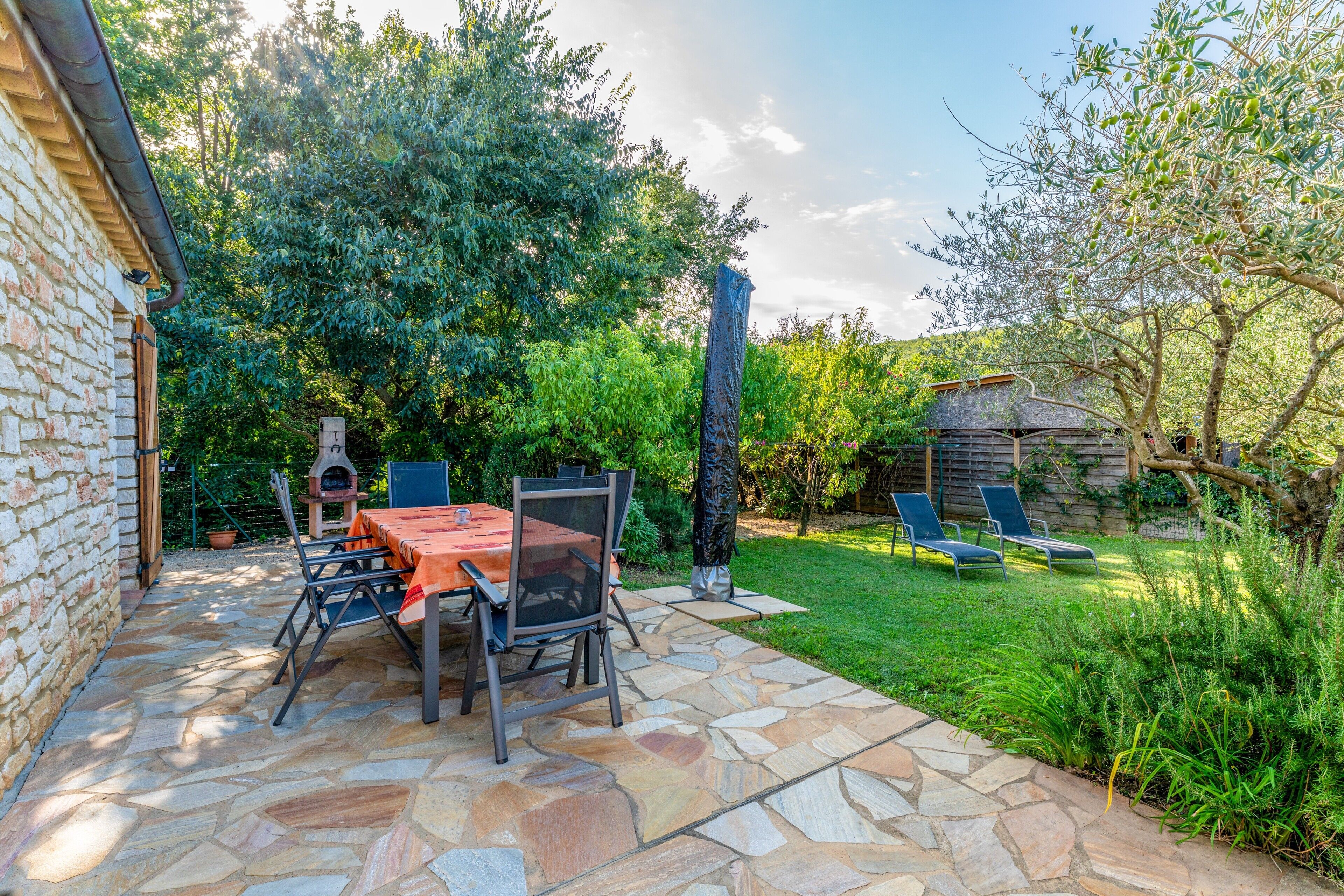 A patio with stone flooring, a table with chairs, a barbecue grill, and a small brick building.