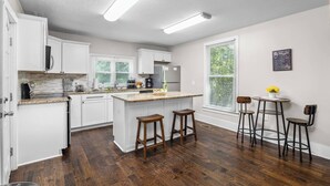 A modern kitchen with white cabinetry, a central island with stools, and a dining area with bar stools. - Masters Classic AUGUSTA (Augusta)