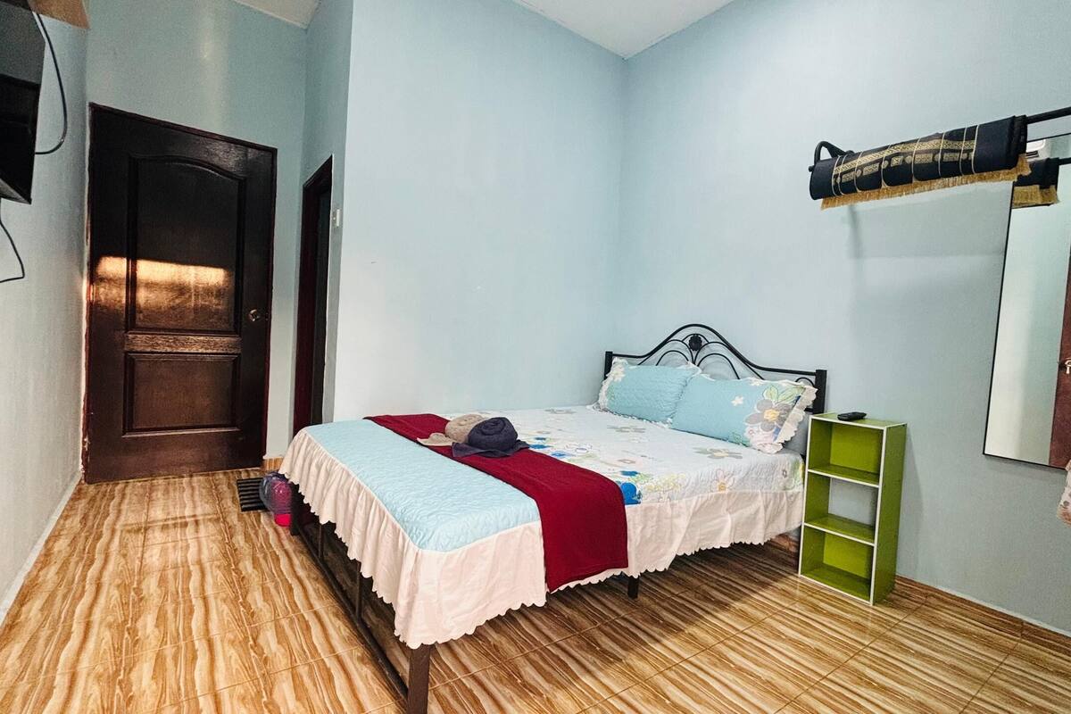 Un dormitorio con cama, suelo de madera, mesita de noche verde, espejo y puerta.