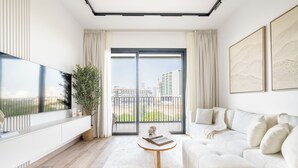 A modern living room with a large TV, a white sofa, a coffee table, and a view of the city through large windows. - JVC Gem 2BR w maidsroom - Oakley Square (Dubai)