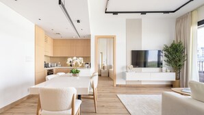 A modern living room with a large flat-screen TV, a dining area with a white table and chairs, and a kitchen with wooden cabinets and a large window. - JVC Gem 2BR w maidsroom - Oakley Square (Dubai)