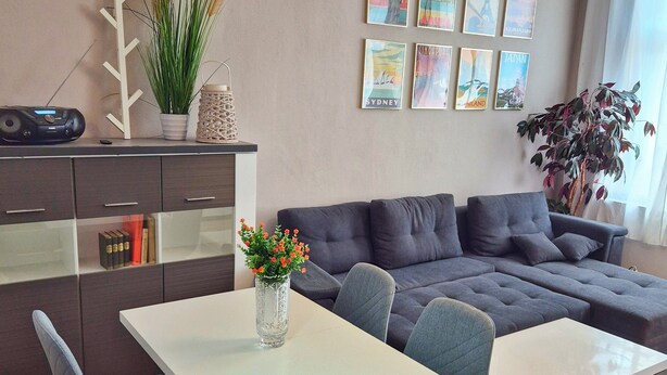 A modern living room with a grey sofa, a white dining table, and chairs. There is a small table with a vase of flowers, a decorative plant, and a wall-mounted speaker.