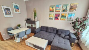 A modern living room with a grey sectional sofa, a white coffee table, and a dining area with a white table and blue chairs. There are travel posters on the wall and a potted plant on the right.