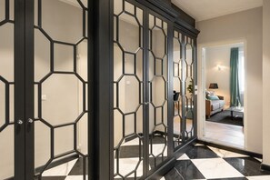 Un couloir avec une porte ornée d’un motif géométrique, un sol à carreaux et, au fond, on aperçoit un salon avec un canapé et une lampe.