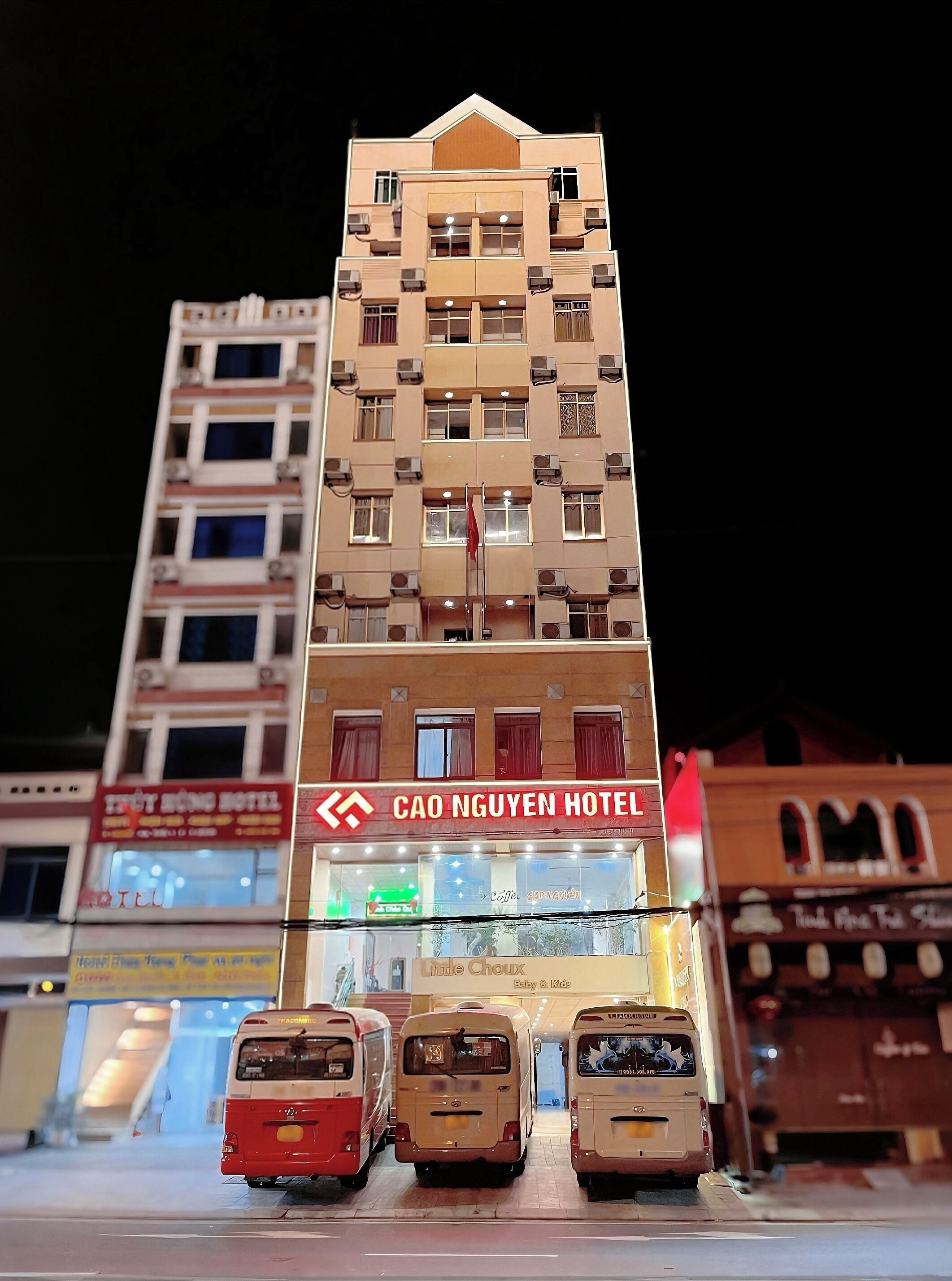 Un immeuble abritant un hôtel, avec plusieurs étages, une enseigne bien visible indiquant « Hôtel Cao Nguyen », et trois autobus stationnés devant.
