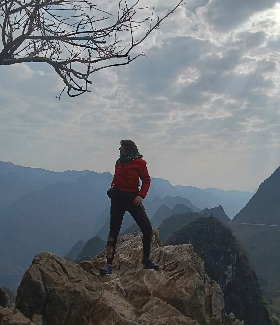 Uma pessoa em um afloramento rochoso, com uma paisagem montanhosa ao fundo.