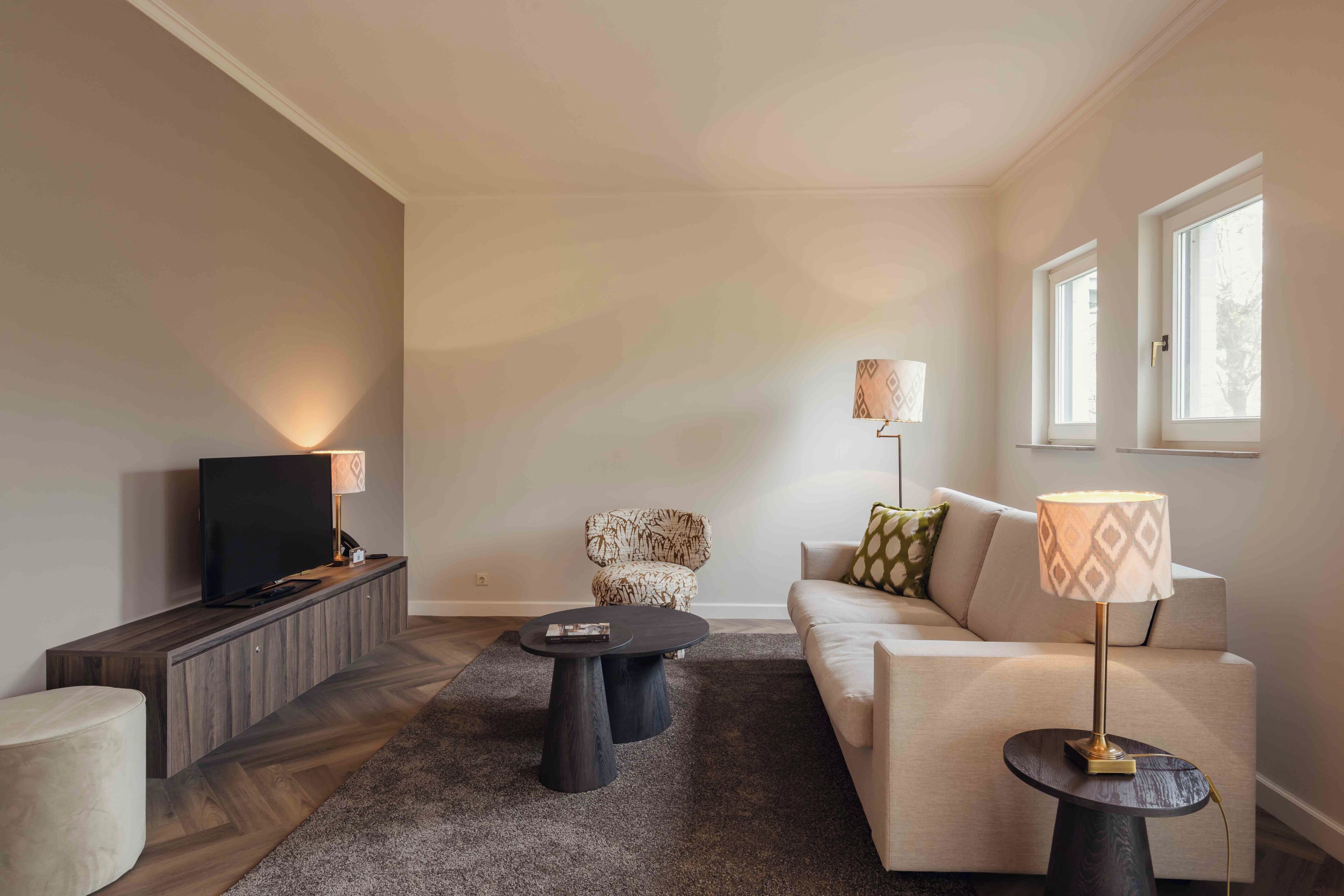A modern living room with a flat-screen TV, two black coffee tables, a patterned armchair, a beige sofa, and a lamp on a side table.