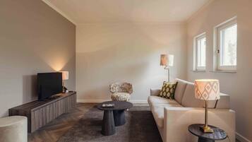 A modern living room with a flat-screen TV, two black coffee tables, a patterned armchair, a beige sofa, and a lamp on a side table.