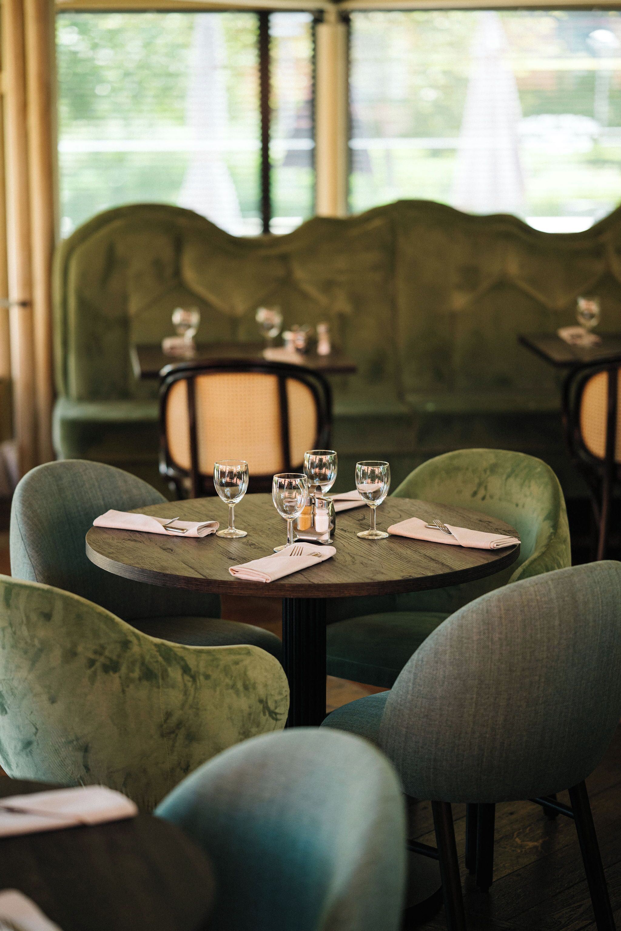Een restaurantinterieur met ronde tafels, groene beklede stoelen en uitzicht op beplanting.