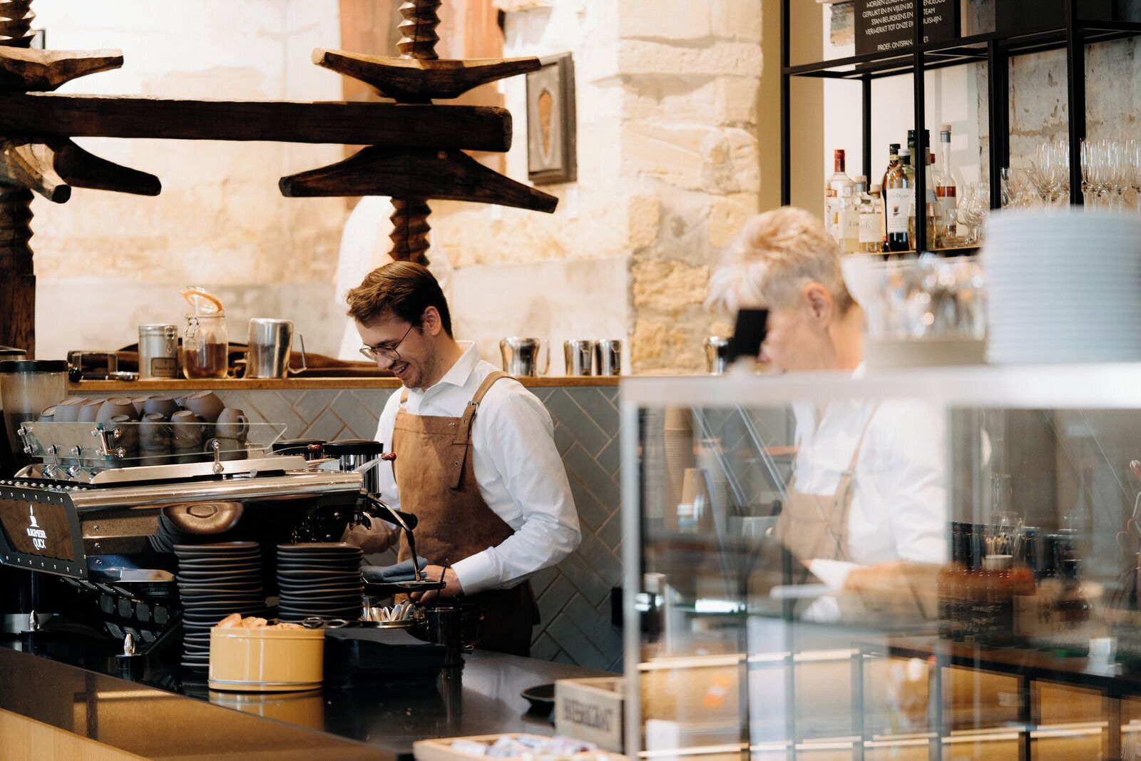 Barista valmistaa kahvia tiskillä, jossa on kahvinkeitin, pino lautasia ja lasivitriini.