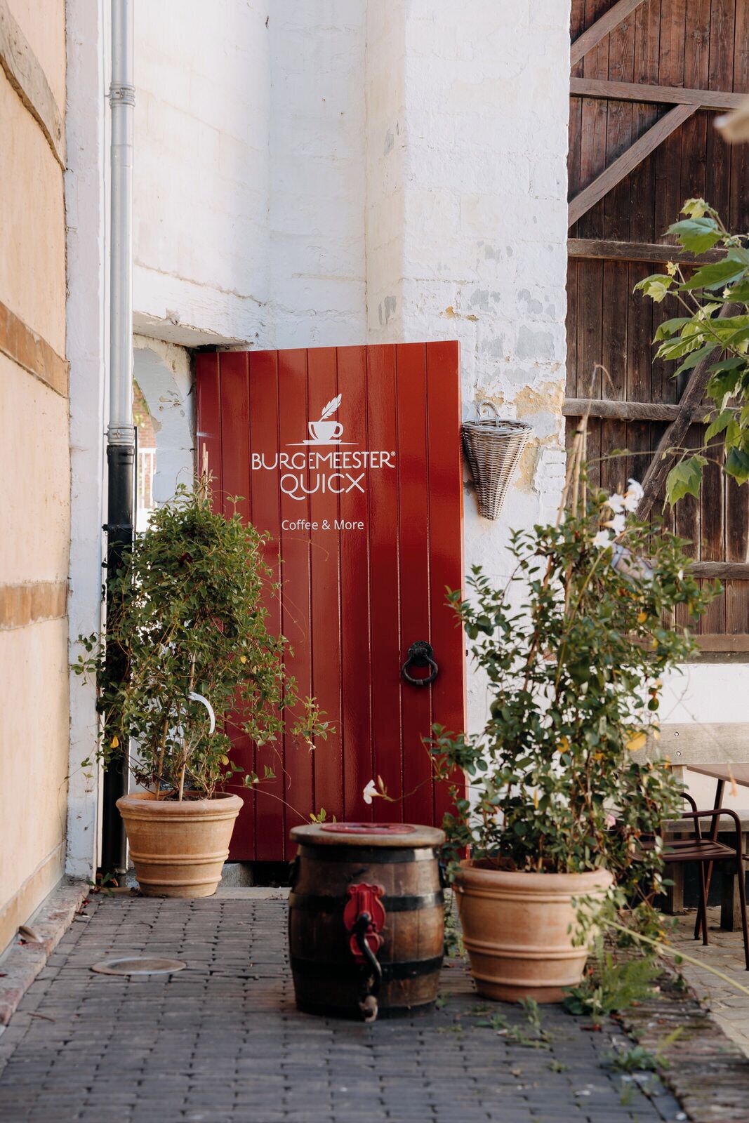 A red door with the text "BURGEMEESTER QUICKX Coffee & More" and two potted plants on either side.