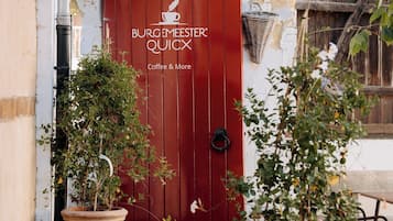 A red door with the text "BURGEMEESTER QUICKX Coffee & More" and two potted plants on either side.