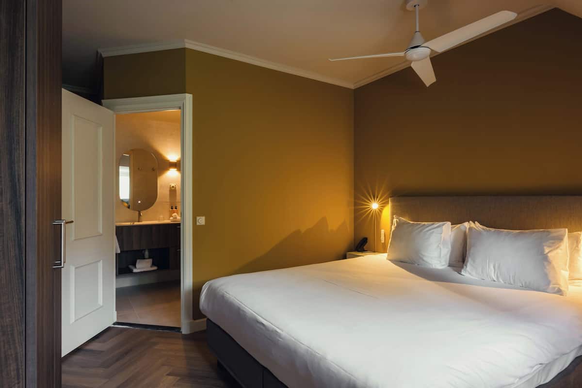 Een hotelkamer met een bed, een plafondventilator, een nachtkastje met een lamp en een zichtbare badkamer door een open deur.