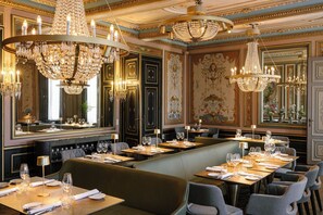 En restaurant med flere spiseborde, elegante lysekroner og detaljerede vægdekorationer.