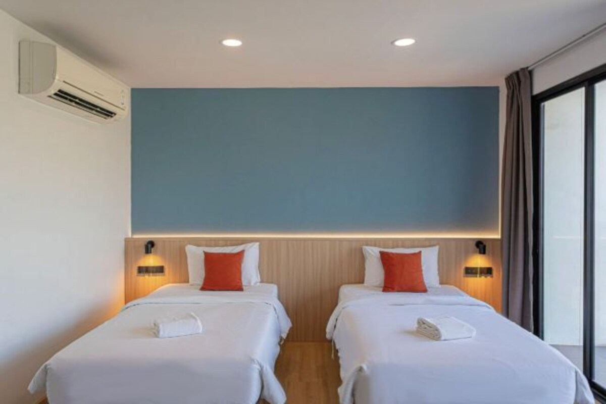 A hotel room with two beds, each with a white bedspread, red pillows, and a white towel. There are wall-mounted lights above each bed and an air conditioning unit on the wall.