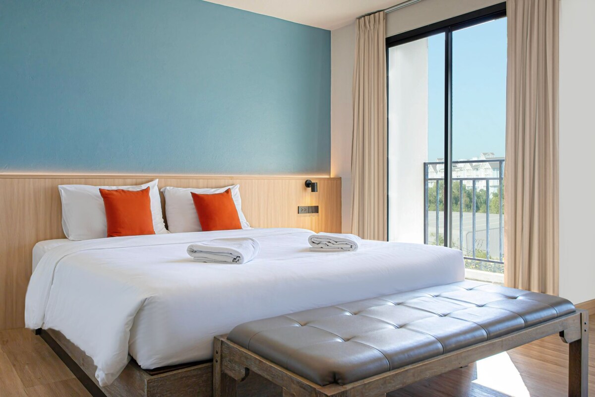 Ein Hotelzimmer mit einem großen Bett, zwei orangefarbenen Kissen, einem Holzkopfende, einer Bank und Blick auf einen Balkon.