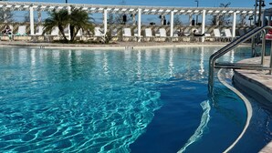 Una piscina cristalina con una pérgola blanca y sillones de descanso al fondo.