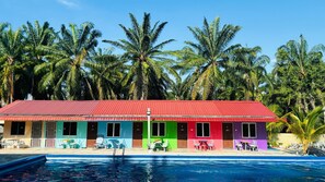 Ein farbenfrohes Gebäude mit rotem Dach und mehreren Zimmern, jedes in einer anderen Farbe. Vor dem Gebäude befindet sich ein Schwimmbecken mit Liegestühlen und Sonnenschirmen.