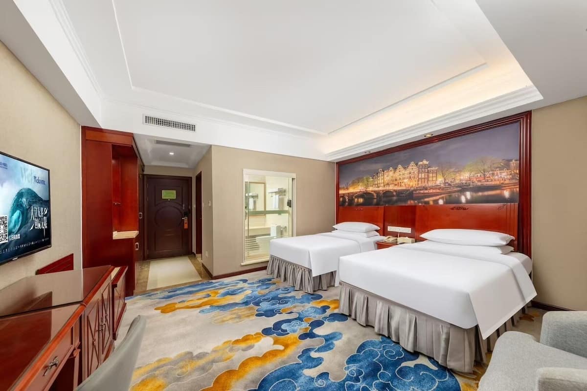 Une chambre d’hôtel avec deux lits, une télévision, un tableau représentant un paysage urbain et une moquette à motifs de nuages.