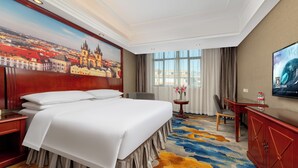 A hotel room with a large bed, a desk, a television, and a painting of a cityscape. - Vienna Hotel International Airport  (Guangzhou)