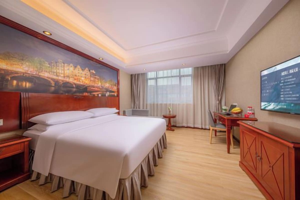 A hotel room with a large bed, a desk with a TV, a painting of a cityscape, and a window with curtains.