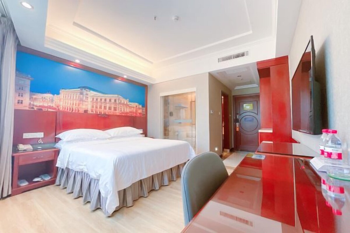 Habitación de hotel con una cama grande, un escritorio, una silla, un televisor y un baño visible a través de una puerta de cristal.