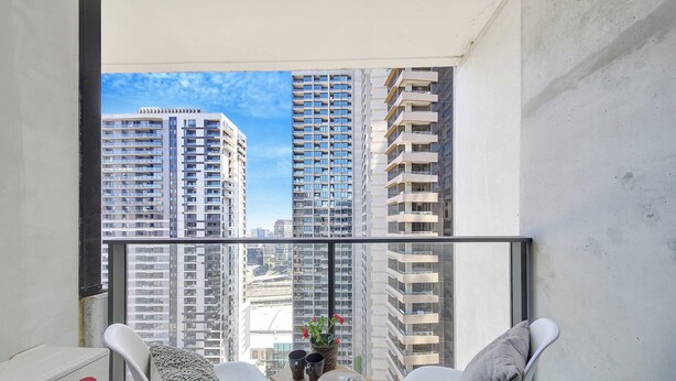 A balcony with two white chairs, a small table with a vase of flowers, and a view of high-rise buildings.