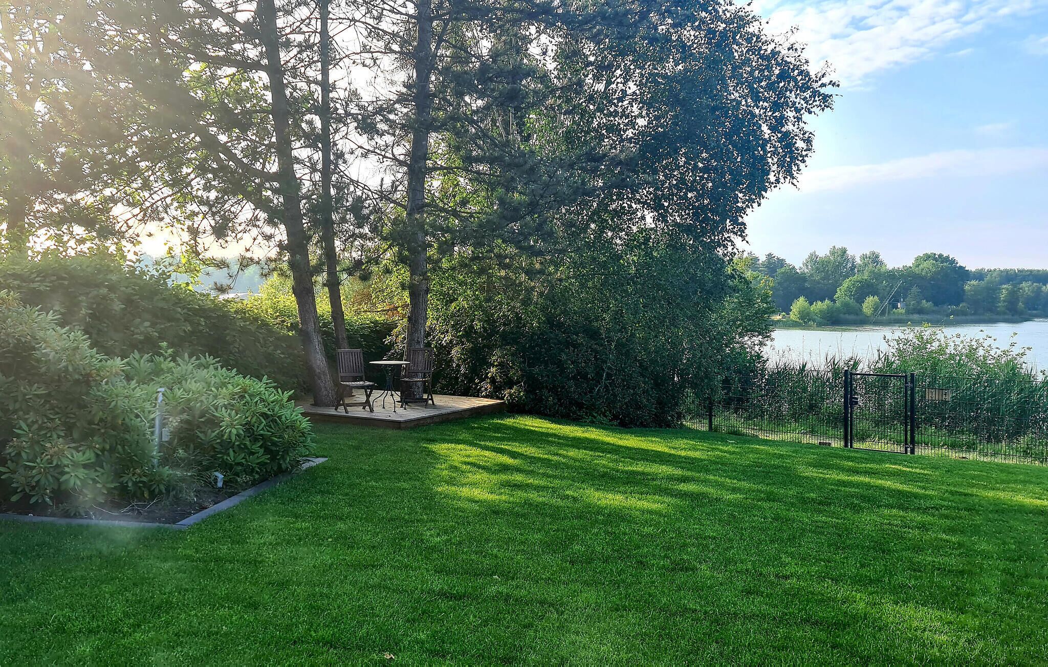 Une pelouse bien entretenue, agrémentée d’une terrasse en bois et de chaises, le tout entouré d’arbres denses et offrant une vue sur un lac.