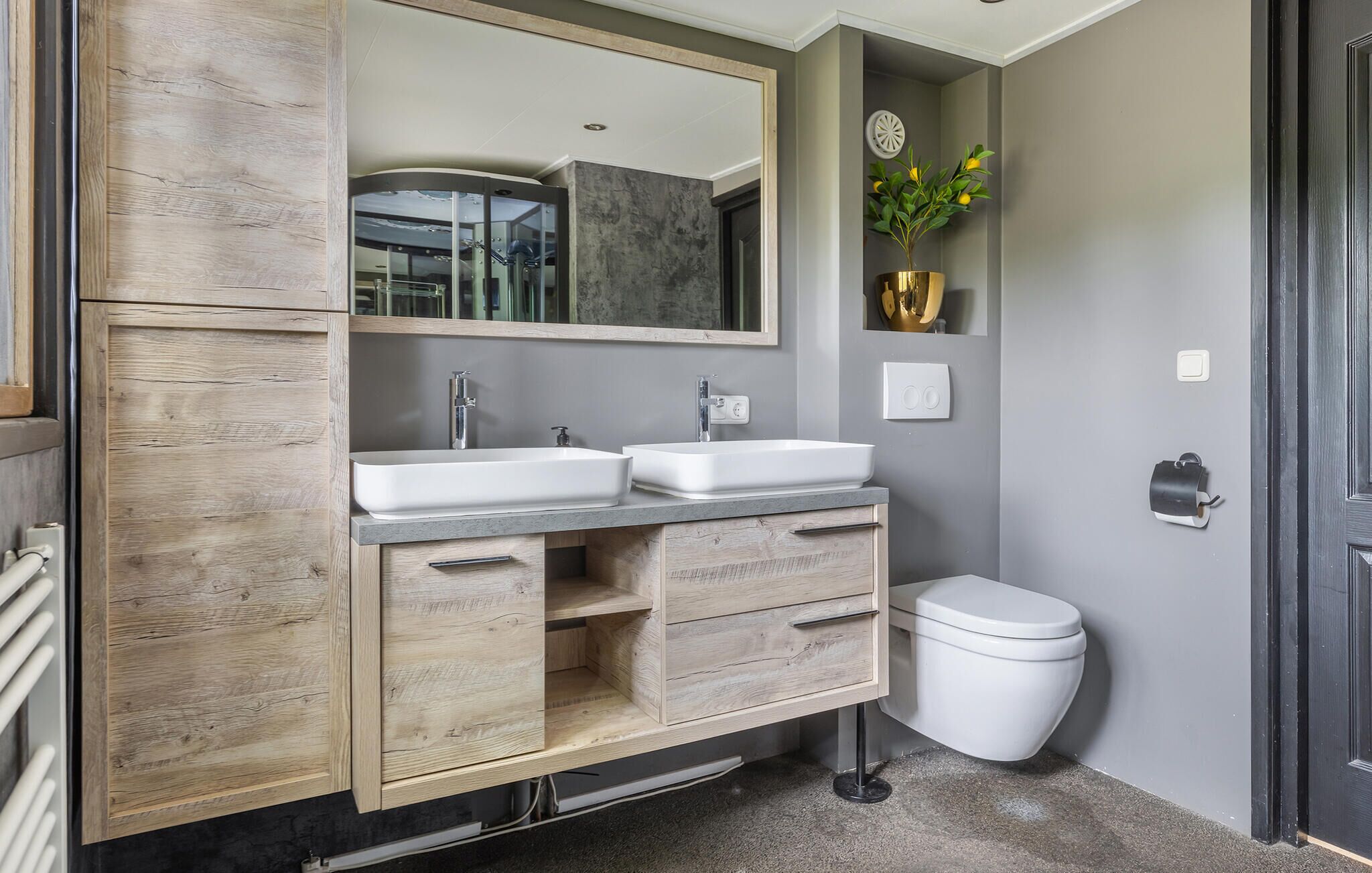 Une salle de bain avec deux lavabos, un miroir, des toilettes et une petite plante accrochée au mur.