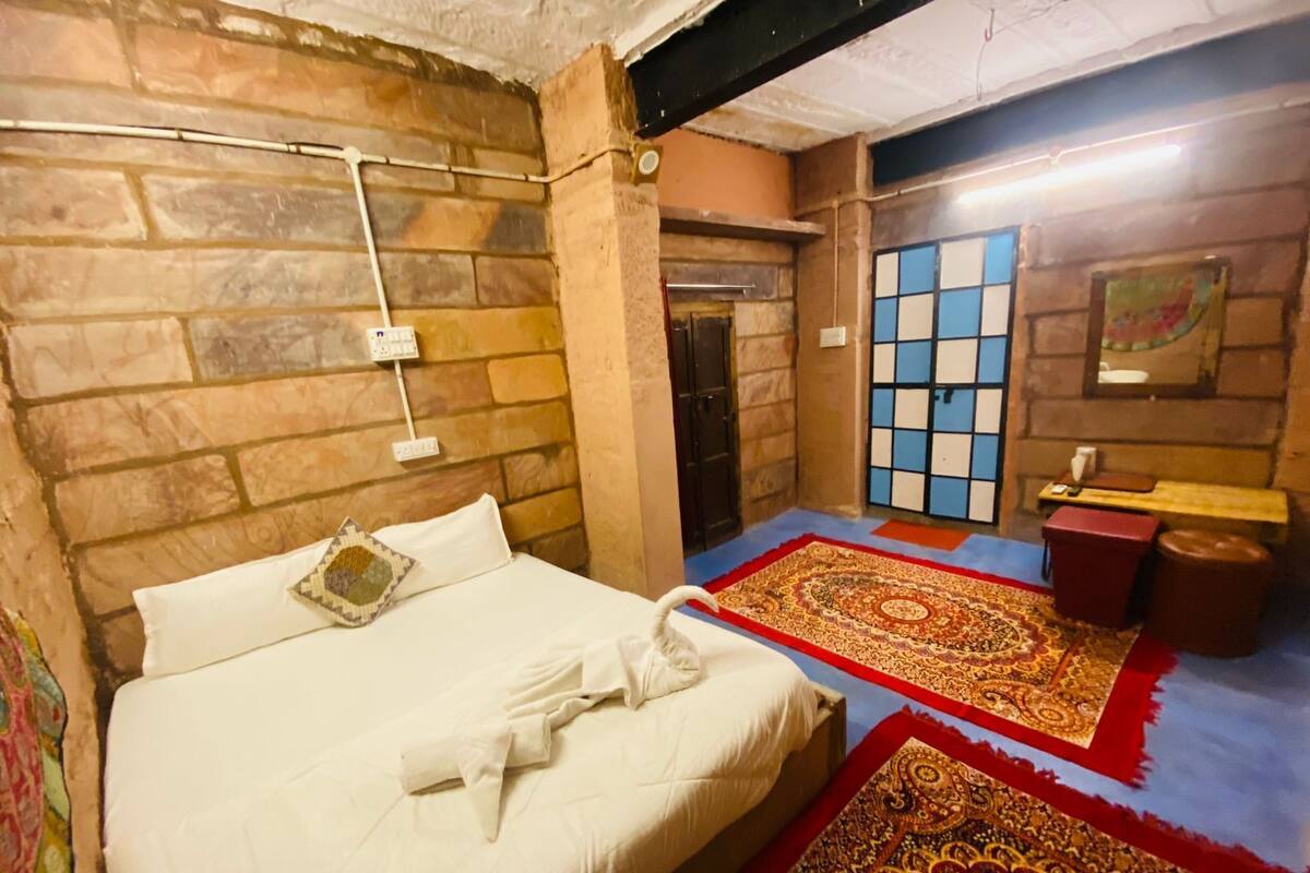 A room with a bed, a blue and red patterned carpet, a small table with a red stool, and a wall with a blue and white grid pattern.