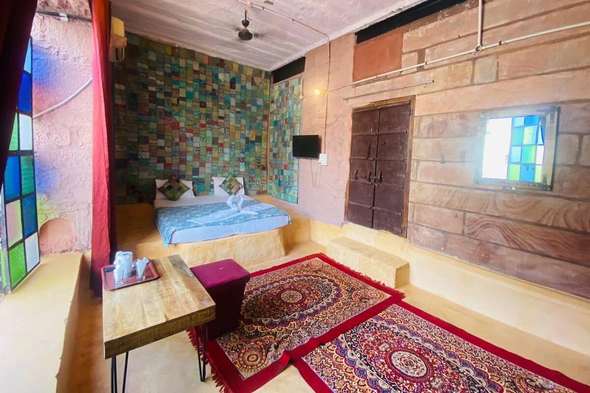 A room with a bed, a table, a red rug, a window, and a wall with a colorful patchwork design.