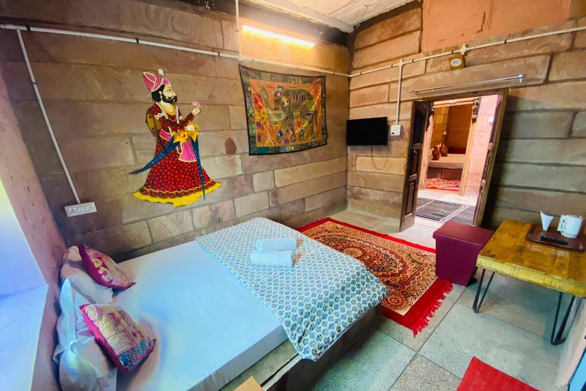 A small, compact room with a bed, a table, and a chair. There is a wall-mounted TV and a door leading to another room.