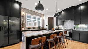 Une cuisine moderne avec des meubles de couleur sombre, des appareils en acier inoxydable, un îlot en marbre avec des tabourets de bar et des luminaires suspendus.