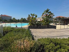 A pool area with a wooden deck, surrounded by greenery and buildings with red roofs.