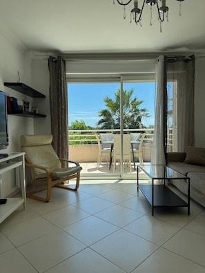 A modern living room with a sofa, a glass coffee table, a TV, and a chair. There is a balcony with a table and chairs, and a view of palm trees.