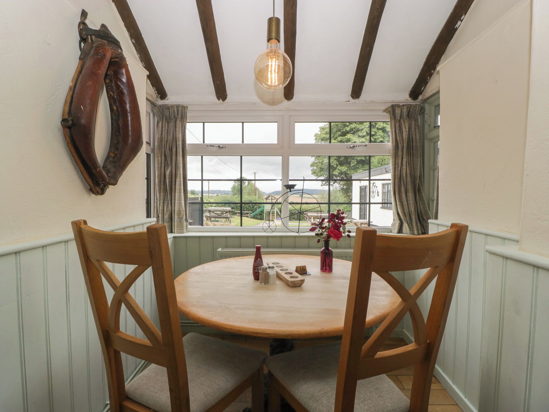 A dining area with a round wooden table, two wooden chairs, a hanging light fixture, and a window with a view of a bicycle and greenery outside.