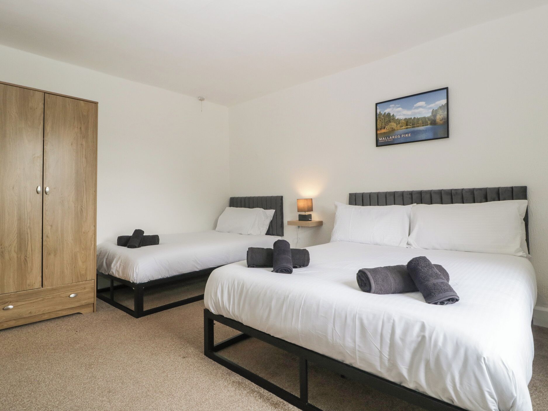 A hotel room with two single beds, a wardrobe, a bedside table with a lamp, and a framed picture on the wall.
