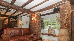Een gezellige woonkamer met een leren bank, een houten bijzettafeltje en rieten fauteuils. Op een raamkozijn staat een grote vaas met gedroogde planten.