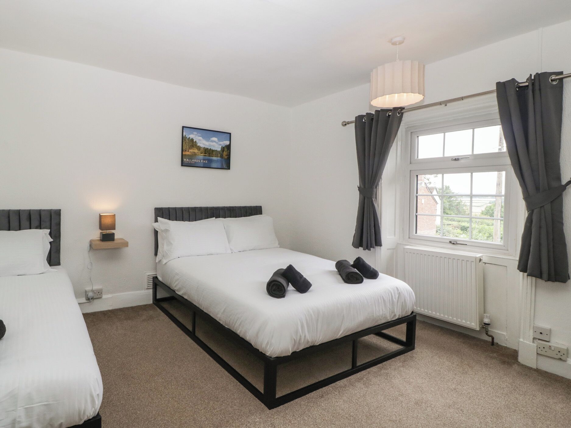 A bedroom with a single bed, a bedside table with a lamp, a window with curtains, and a picture on the wall.