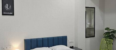 A bedroom with a single bed, a bedside table with a lamp, a green chair, and a framed picture on the wall.
