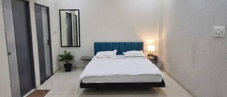 A hotel room with a bed, bedside table, lamp, air conditioning unit, ceiling fan, and a small potted plant.